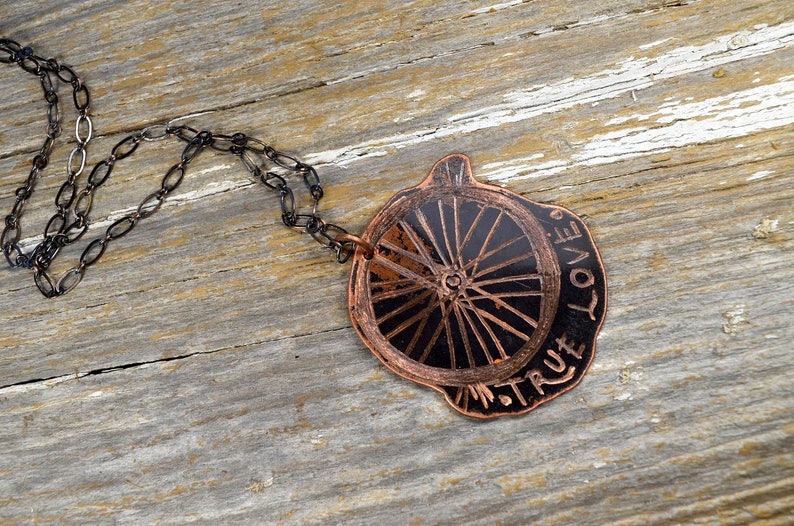 May include: A copper necklace with a black and copper wheel pendant. The pendant is inscribed with the words "True Love".