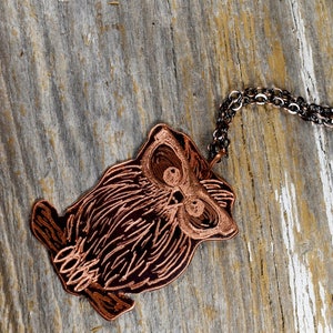 以下が含まれることがあります： 銅色のフクロウペンダントネックレス。フクロウのペンダントは、詳細なエッチングラインで装飾され、暗いチェーンから吊り下げられています。 ネックレスは、風化した木製の表面に表示されています。