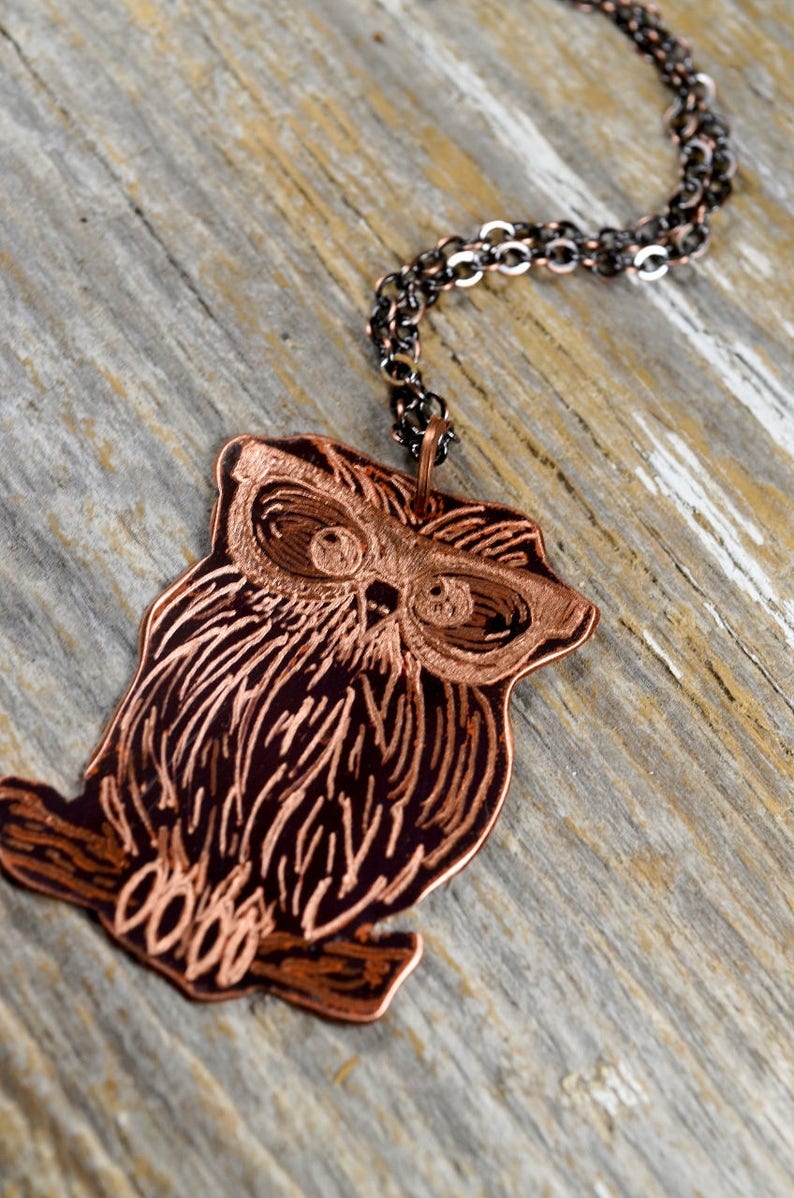 以下が含まれることがあります： 銅色のフクロウペンダントネックレス。ペンダントは、詳細な羽と目のマーキングが施されたエッチングされたフクロウのデザインが特徴です。ネックレスは濃い色のチェーンで、風化した木製の表面に表示されています。