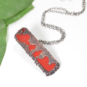 May include: A red and copper necklace with a rectangular pendant featuring a textured surface and three red heart shapes. The pendant is attached to a silver chain.