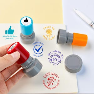 Könnte beinhalten: Eine Sammlung farbenfroher Lehrerstempel mit verschiedenen Designs und Phrasen, darunter ein Daumen hoch, eine Sonnenblume und die Worte „Terrific!“ und „Good Work!“. Die Stempel sind rot, blau und orange.