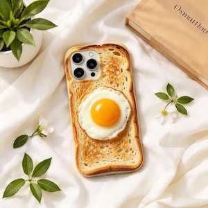 Peut inclure: Coque de téléphone représentant une tranche de pain grillé surmontée d'un œuf au plat. Le boîtier a une bordure transparente. L'image est posée sur un fond de tissu blanc avec de la verdure et un livre.