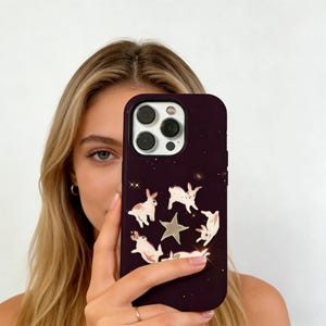 以下が含まれることがあります： 白いウサギ、銀色の星、小さな白い星のデザインが施された濃い紫色の携帯電話ケース。ケースは光沢仕上げで、カメラの切り抜きがあります。