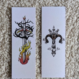 Peut inclure: Deux marque-pages rectangulaires blancs. L'un présente un dessin d'épée avec des flammes et des vignes épineuses. L'autre marque-page présente un dessin de croix avec des cornes de bélier et des motifs floraux. Les dessins sont en noir, gris, rouge et jaune.