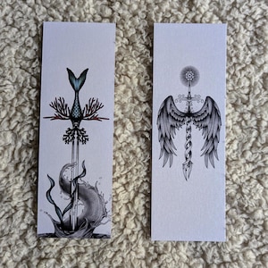 Peut inclure: Deux marque-pages rectangulaires blancs avec des illustrations détaillées en noir et bleu. L'un représente une épée avec une queue de sirène et du corail. L'autre montre une épée avec des ailes d'ange et une étoile.