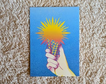 May include: A postcard featuring a hand holding an ice cream cone with a sunburst design. The cone is yellow and the sun is yellow and orange against a blue background. The hand is a light skin tone.