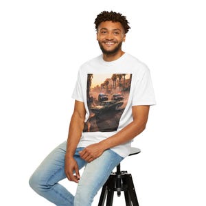 Puede incluir: Camiseta blanca con un gráfico que representa una escena de persecución de coches con coches de policía y palmeras, con un fondo de puesta de sol. La persona lleva la camiseta y está sentada en un taburete negro. La imagen tiene un ambiente cinematográfico.