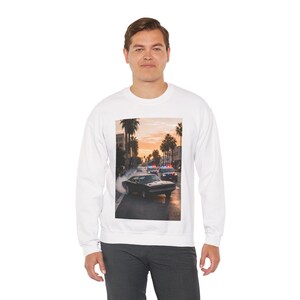 Puede incluir: Sudadera blanca de manga larga con un estampado gráfico de un coche negro derrapando en una calle de la ciudad con palmeras y coches de policía. La imagen tiene un fondo de puesta de sol.