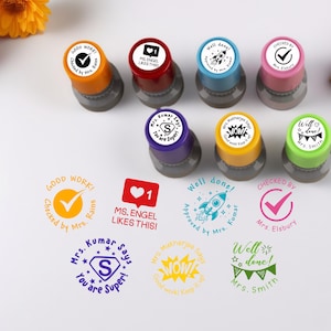 Pode incluir: Uma coleção de carimbos auto-entintados coloridos com vários desenhos e texto, incluindo "Good Work!", um coração com "MS. ENGEL LIKES THIS!" e um foguete com "Well done! Approved by Mrs. Kumar".