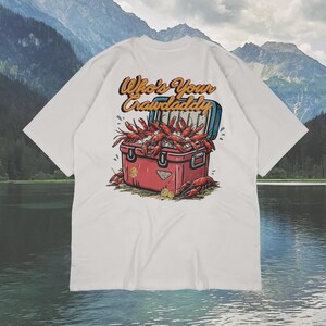 以下が含まれることがあります： 白いTシャツ。赤いクーラーボックスからザリガニがあふれ出ているグラフィック。クーラーボックスは開いており、ザリガニがこぼれ落ちています。「Who's Your Crawdaddy」の文字がレトロなフォントでクーラーボックスの上にアーチ状に配置されています。背景には湖と山が描かれています。