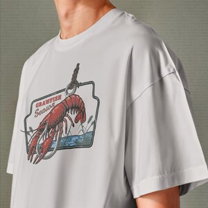 Pu&ograve; includere: T-shirt grigio chiaro con una grafica di gambero rosso e la scritta "Crawfish Season". Il design include un amo da pesca, acqua e canne, in stile vintage.