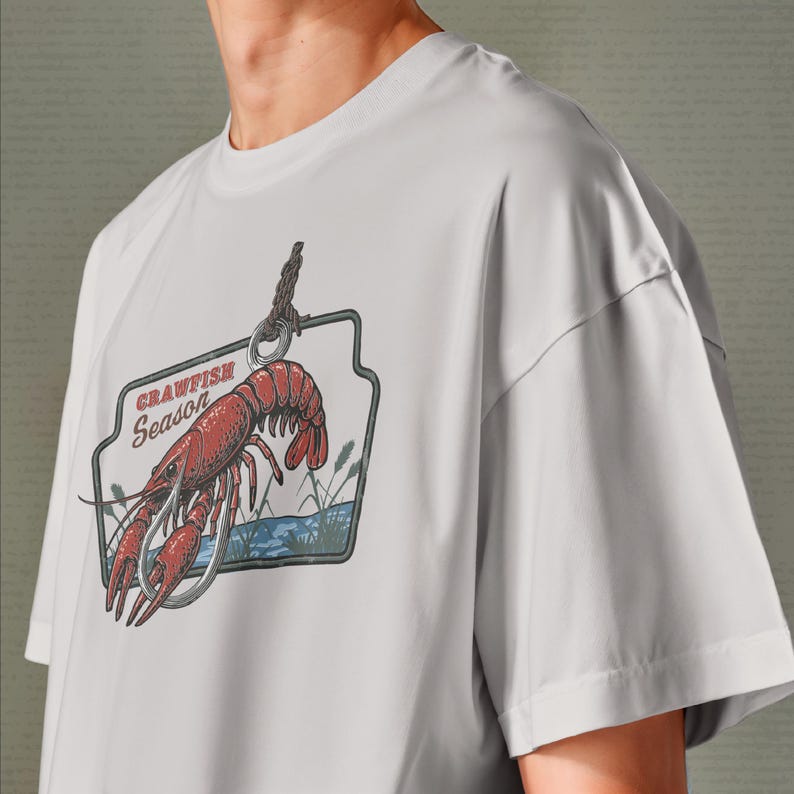 Pu&ograve; includere: T-shirt grigio chiaro con una grafica di gambero rosso e la scritta "Crawfish Season". Il design include un amo da pesca, acqua e canne, in stile vintage.