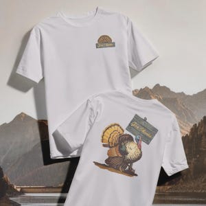 May include: Two white t-shirts. One shirt has a small graphic on the front with the text "Give Thanks". The other shirt has a large turkey graphic on the back, holding a sign that says "Give Thanks".