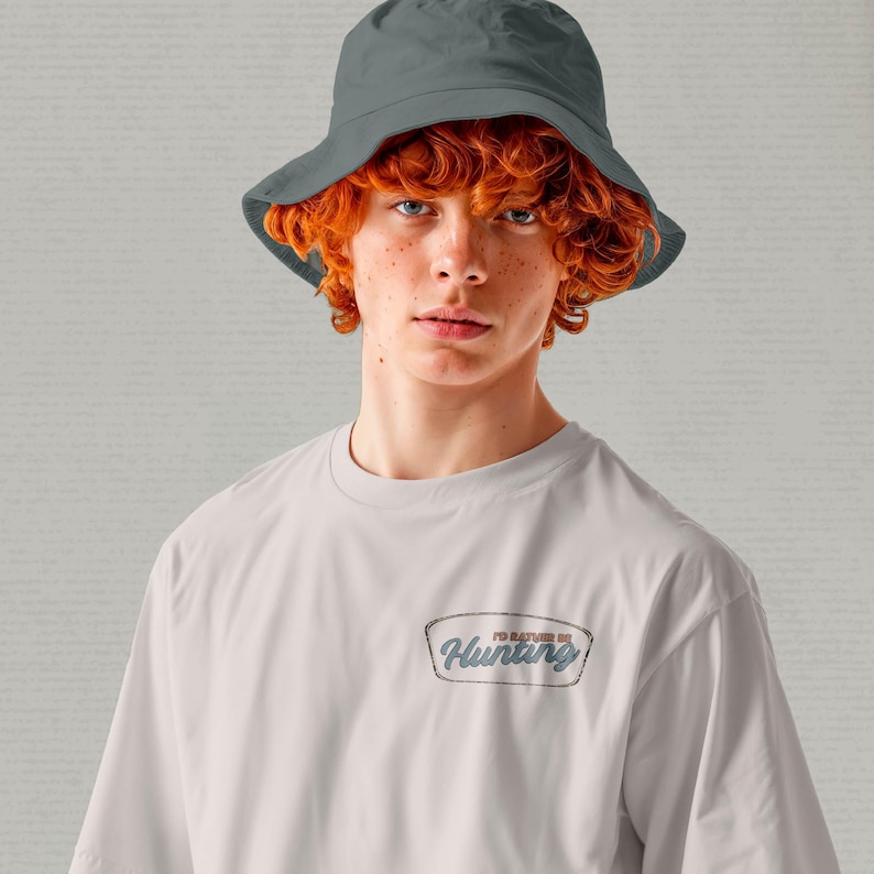 May include: A person wearing a gray bucket hat and a white t-shirt. The t-shirt has a rectangular patch with the text "I'd Rather Be Hunting" in a blue and gray design.
