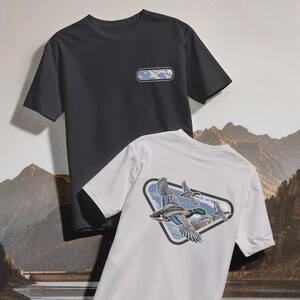 May include: Two short-sleeved t-shirts. One is charcoal gray with a rectangular patch on the chest. The other is white, featuring a triangular graphic of flying ducks with a mountain and sky backdrop. The shirts are made of a soft material.