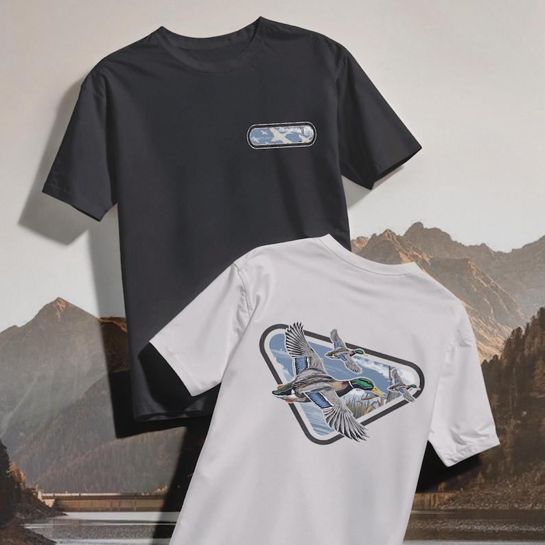 May include: Two short-sleeved t-shirts. One is charcoal gray with a rectangular patch on the chest. The other is white, featuring a triangular graphic of flying ducks with a mountain and sky backdrop. The shirts are made of a soft material.