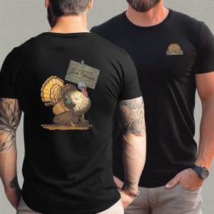 May include: Black t-shirt featuring a colorful turkey illustration holding a wooden sign that reads "Give Thanks." The front of the shirt has a small sunburst design. A Thanksgiving-themed graphic tee.