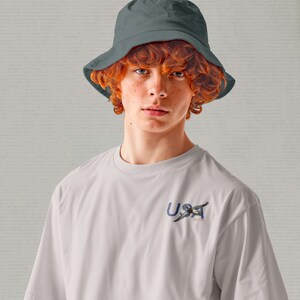 May include: A person wearing a gray bucket hat and a white t-shirt. The t-shirt has a small graphic with the word "USA" and a bird design. The person has red curly hair and is looking at the camera.
