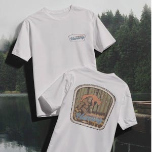 May include: Two white t-shirts. One shirt has a small patch with the word "Hunting" in a blue and tan design. The other shirt has a large graphic on the back featuring a Bigfoot and a deer in a forest setting, with the word "Hunting" below.