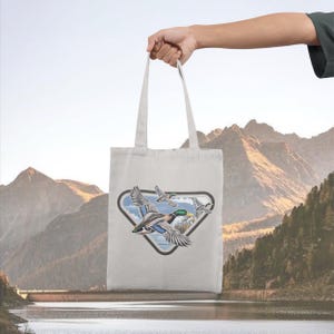 May include: A light-colored tote bag featuring a graphic of flying ducks within a triangular frame. The ducks are depicted with detailed feathers and vibrant colors. The bag is held up against a backdrop of mountains and a lake.