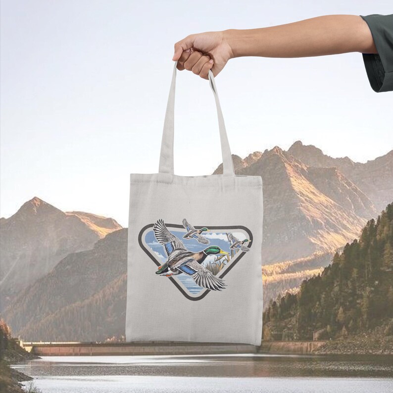 May include: A light-colored tote bag featuring a graphic of flying ducks within a triangular frame. The ducks are depicted with detailed feathers and vibrant colors. The bag is held up against a backdrop of mountains and a lake.