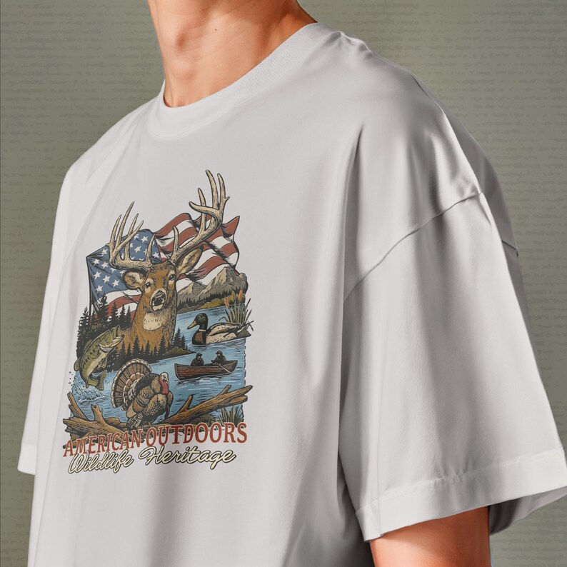 以下が含まれることがあります： 白いTシャツ。鹿、アメリカ国旗、山、アヒルとボートのある湖、七面鳥、魚の詳細なグラフィックが特徴です。「AMERICAN OUTDOORS Wildlife Heritage」のテキストが下にあります。