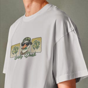 May include: A light gray t-shirt featuring a graphic of a duck wearing sunglasses, a camouflage hat, and a gold chain. The duck is holding a shamrock in its beak, with the words "Lucky Duck" below the image.