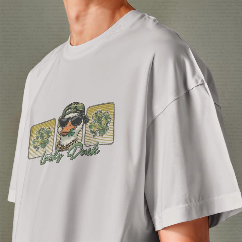 May include: A light gray t-shirt featuring a graphic of a duck wearing sunglasses, a camouflage hat, and a gold chain. The duck is holding a shamrock in its beak, with the words "Lucky Duck" below the image.