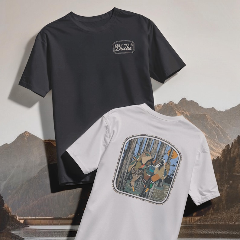 Op de afbeelding: Twee T-shirts met korte mouwen. E&eacute;n is antracietgrijs met "KEEP YOUR DUCKS" op de borst gedrukt. De andere is wit, met een afbeelding van eenden in een bosrijk gebied op de achterkant. De achtergrond toont een bergketen.