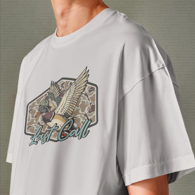May include: A light gray t-shirt featuring a graphic of a duck with outstretched wings inside a brown and gray camouflage hexagon. The text "Last Call" is written in a script font below the duck.