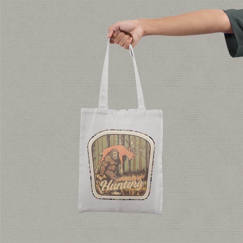May include: A white tote bag featuring a graphic of a Bigfoot-like creature and a deer in a forest setting. The text "I'd Rather Be Hunting" is below the image. The bag is held by a person.