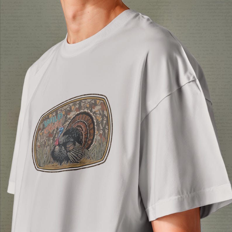 May include: A light gray t-shirt featuring a detailed illustration of a wild turkey within a rectangular frame. The turkey is depicted with brown, black, and blue feathers, set against a backdrop of autumn foliage and the text "WILD GAME".