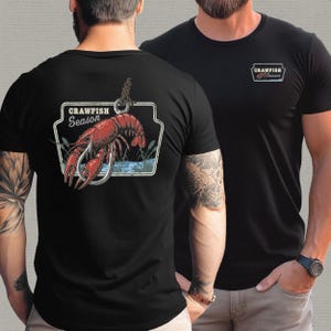 Pu&ograve; includere: T-shirt nere con un design grafico raffigurante un gambero rosso infilzato su un amo da pesca, con la scritta "CRAWFISH Season". Il disegno &egrave; sul retro e una versione pi&ugrave; piccola sul petto sinistro.
