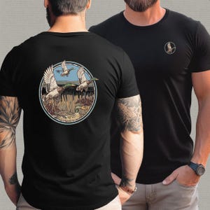 Może przedstawiać: Czarny t-shirt z kr&oacute;tkim rękawem z okrągłą grafiką przedstawiającą kaczki w locie nad bagnem. Projekt zawiera kaczki, trzciny i błękitne niebo. Mniejsza wersja grafiki znajduje się na lewej piersi. Koszulka wykonana jest z miękkiego materiału.