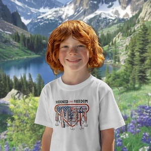 May include: A white t-shirt with the words "HOOKED ON FREEDOM" and a graphic of fishing lures in an American flag design. The shirt is worn by a person with red hair, smiling against a backdrop of mountains and a lake.