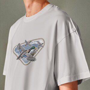 May include: A light gray t-shirt featuring a graphic of three flying ducks. The ducks are depicted with detailed feathers, green heads, and yellow beaks, set against a backdrop of water and reeds within a diamond-shaped frame.