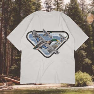 May include: White t-shirt featuring a graphic of three flying ducks within a black-outlined, triangular shape. The ducks have detailed plumage in shades of gray, blue, green, and brown. The background shows a landscape with water and trees.