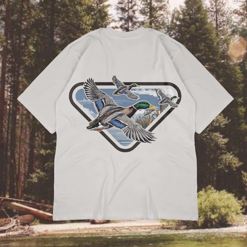 May include: White t-shirt featuring a graphic of three flying ducks within a black-outlined, triangular shape. The ducks have detailed plumage in shades of gray, blue, green, and brown. The background shows a landscape with water and trees.