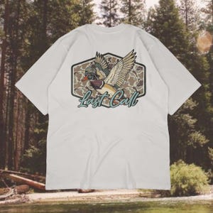 May include: White t-shirt featuring a detailed graphic of a duck in flight, wearing a hat and goggles, set against a camouflage background. The text "Last Call" is written in a teal script below the duck. The shirt has short sleeves.
