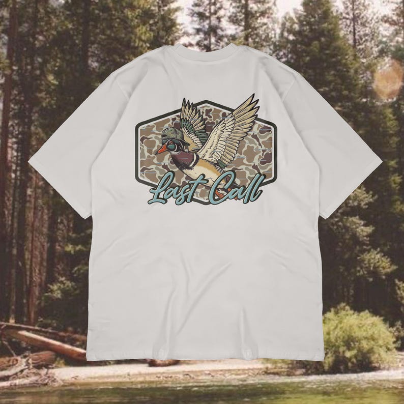 May include: White t-shirt featuring a detailed graphic of a duck in flight, wearing a hat and goggles, set against a camouflage background. The text "Last Call" is written in a teal script below the duck. The shirt has short sleeves.