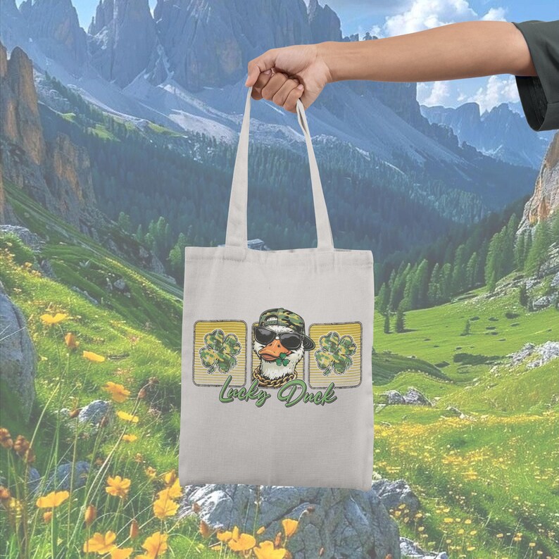 May include: A white tote bag featuring a graphic of a duck wearing sunglasses, a camouflage hat, and a gold chain. The design includes shamrocks and the text "Lucky Duck" in green. The bag is held up against a scenic mountain backdrop.