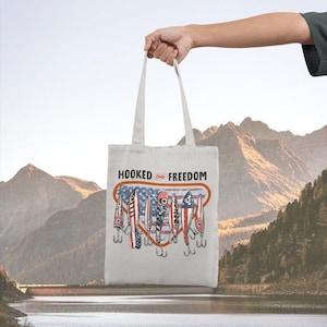 May include: A cream-coloured tote bag featuring the text "HOOKED ON FREEDOM" above a graphic of fishing lures with American flag designs. The bag is held up against a backdrop of mountains and water.