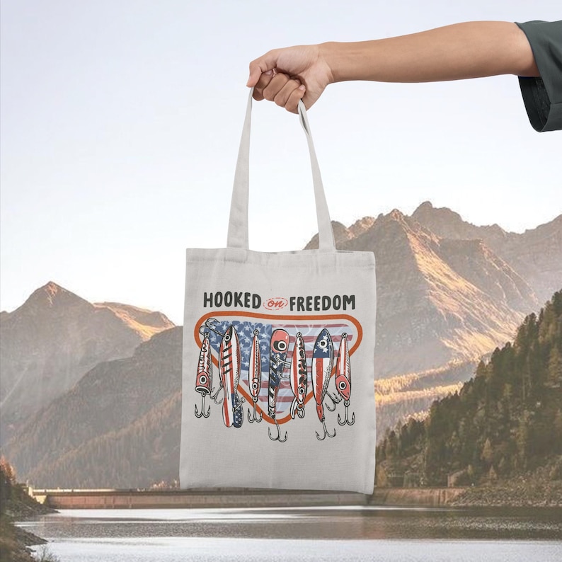 May include: A cream-coloured tote bag featuring the text "HOOKED ON FREEDOM" above a graphic of fishing lures with American flag designs. The bag is held up against a backdrop of mountains and water.