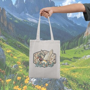 May include: A cream-colored tote bag with a graphic of a duck wearing a pilot's hat and wings, set against a camouflage background. The text "Last Call" is below the graphic. The bag is held up against a backdrop of mountains and green fields.
