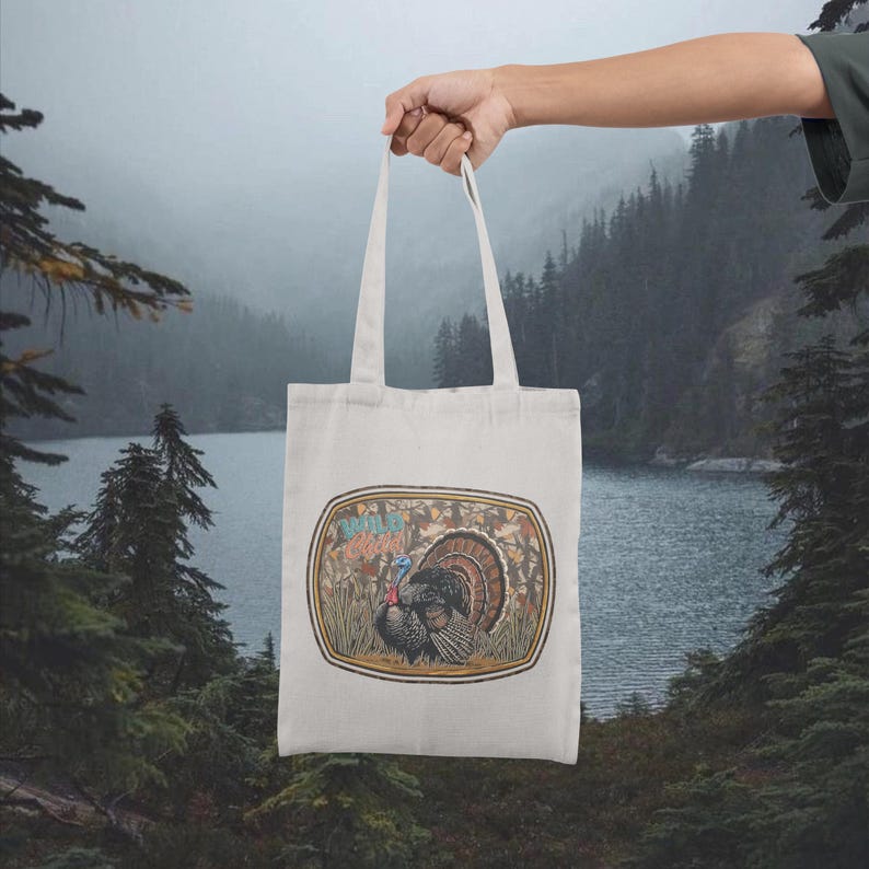 May include: Off-white tote bag featuring a colorful illustration of a turkey with the text "Wild Turkey" in a vintage style. The bag is held up against a backdrop of a lake and forest.