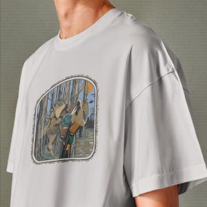 Op de afbeelding: Lichtgrijs t-shirt met een natuursc&egrave;ne met eenden die aan een boom hangen. De afbeelding is omlijst met een decoratieve rand. Het shirt heeft korte mouwen en een ronde hals.