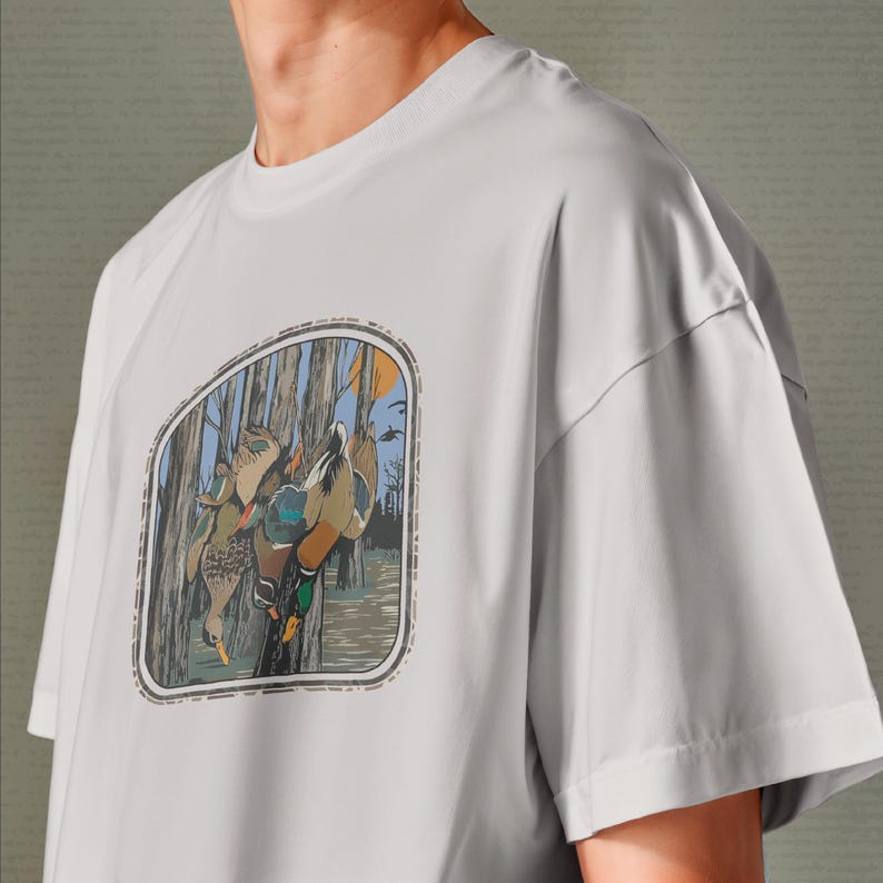 Op de afbeelding: Lichtgrijs t-shirt met een natuursc&egrave;ne met eenden die aan een boom hangen. De afbeelding is omlijst met een decoratieve rand. Het shirt heeft korte mouwen en een ronde hals.