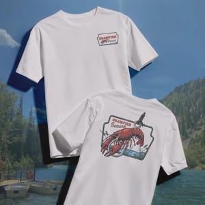 Pu&ograve; includere: Due magliette bianche. Una ha una piccola grafica rettangolare sul davanti con "Crawfish Season". L'altra ha una grafica pi&ugrave; grande sul retro con un gambero rosso, un amo e le parole "Crawfish Season".