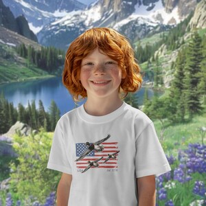 May include: White t-shirt featuring a graphic of flying ducks over a stylized American flag. The shirt includes the text "USA" and "Est. 1776". The background shows a scenic mountain landscape with a lake and trees.