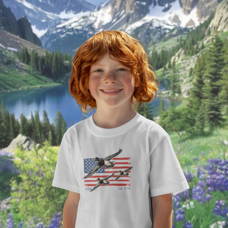 May include: White t-shirt featuring a graphic of flying ducks over a stylized American flag. The shirt includes the text "USA" and "Est. 1776". The background shows a scenic mountain landscape with a lake and trees.